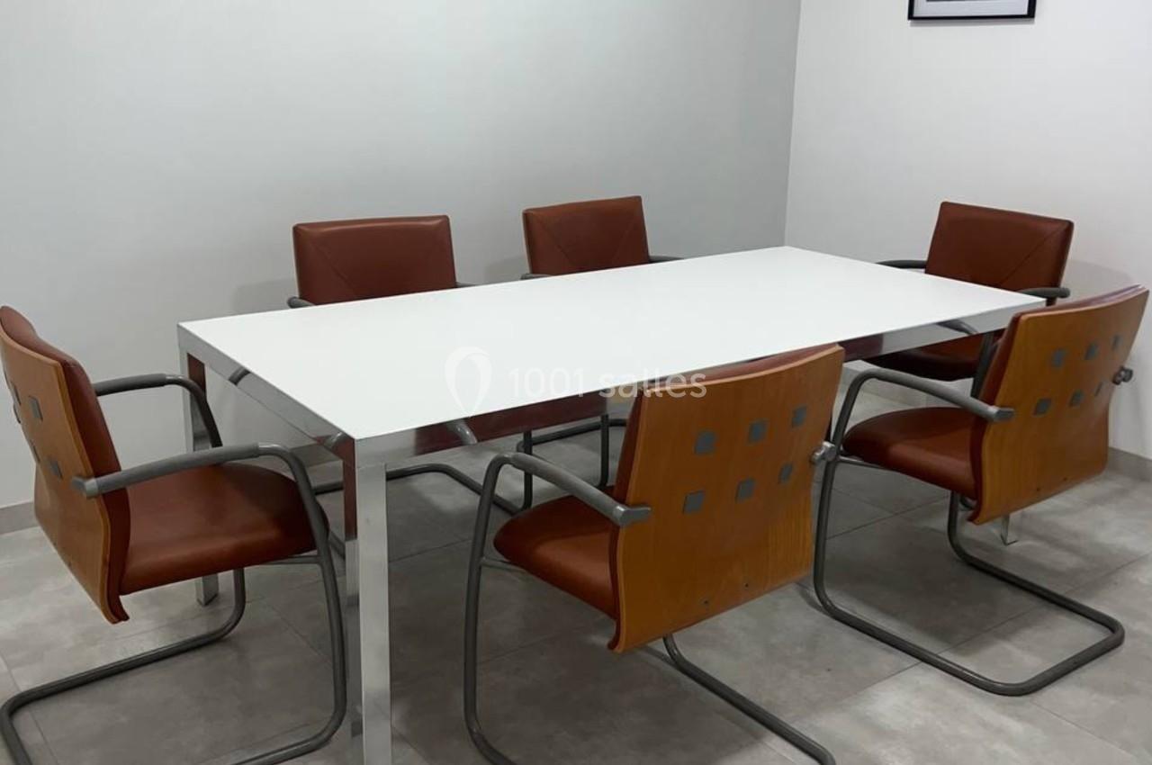 Salle de réunion avec une table blanche rectangulaire entourée de six chaises en cuir marron sur un sol carrelé.