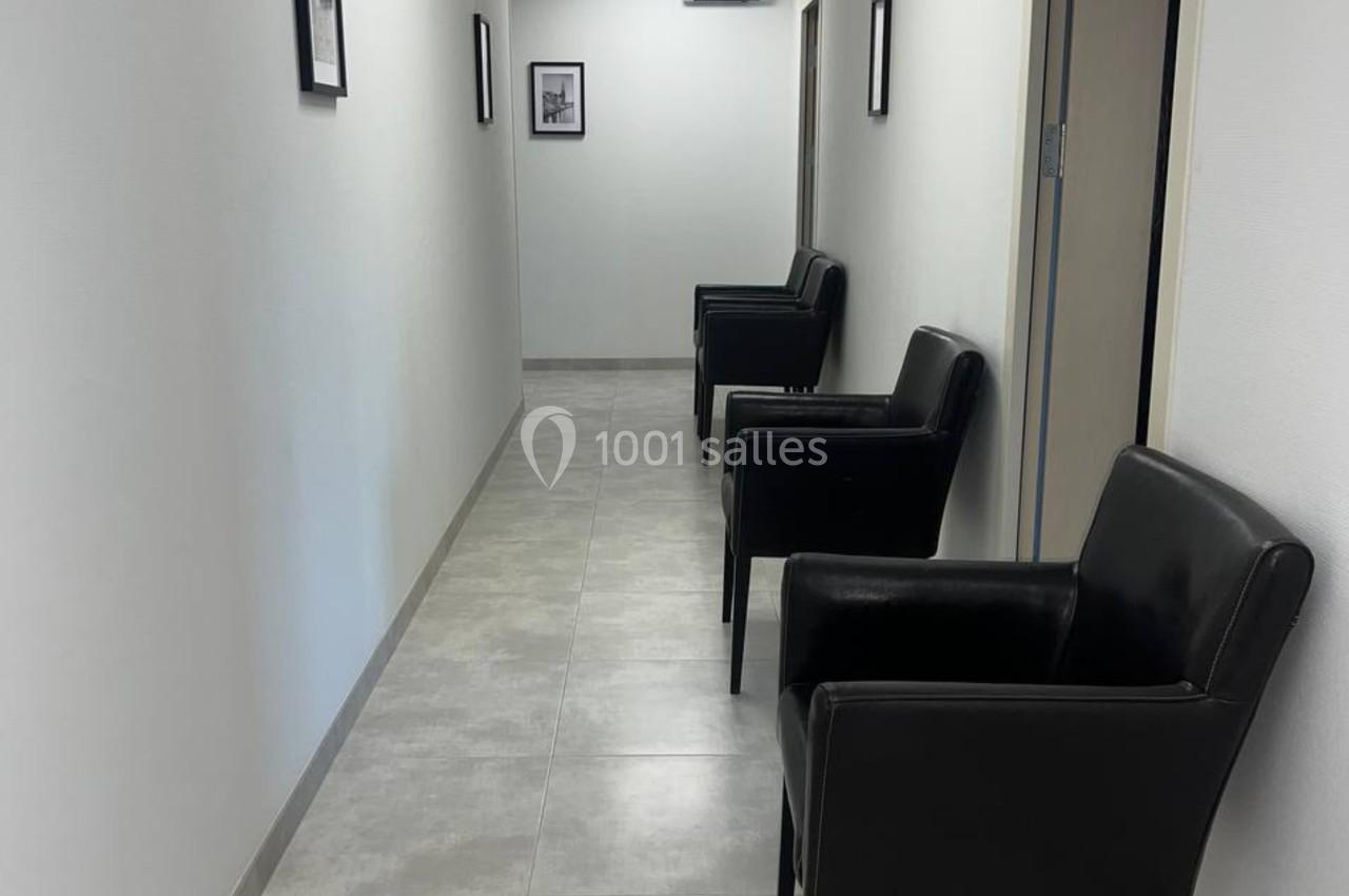 Couloir lumineux avec sol en carrelage, murs blancs, trois fauteuils noirs alignés et cadres accrochés aux murs.