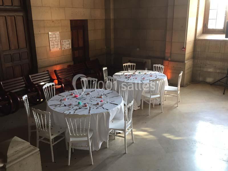 Salle lumineuse avec deux tables rondes dressées, entourées de chaises blanches, près de bancs en bois et d'une fenêtre.