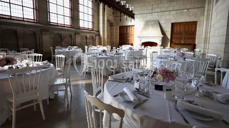 Salle de réception élégante avec tables rondes dressées, chaises blanches et grandes fenêtres laissant entrer la lumière.