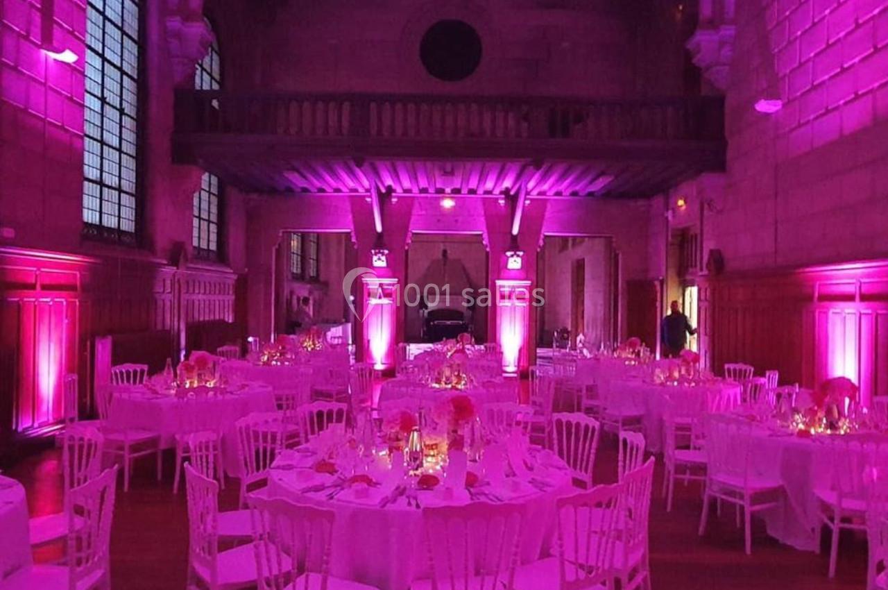 Salle de réception éclairée en rose avec des tables rondes dressées et des chaises blanches.