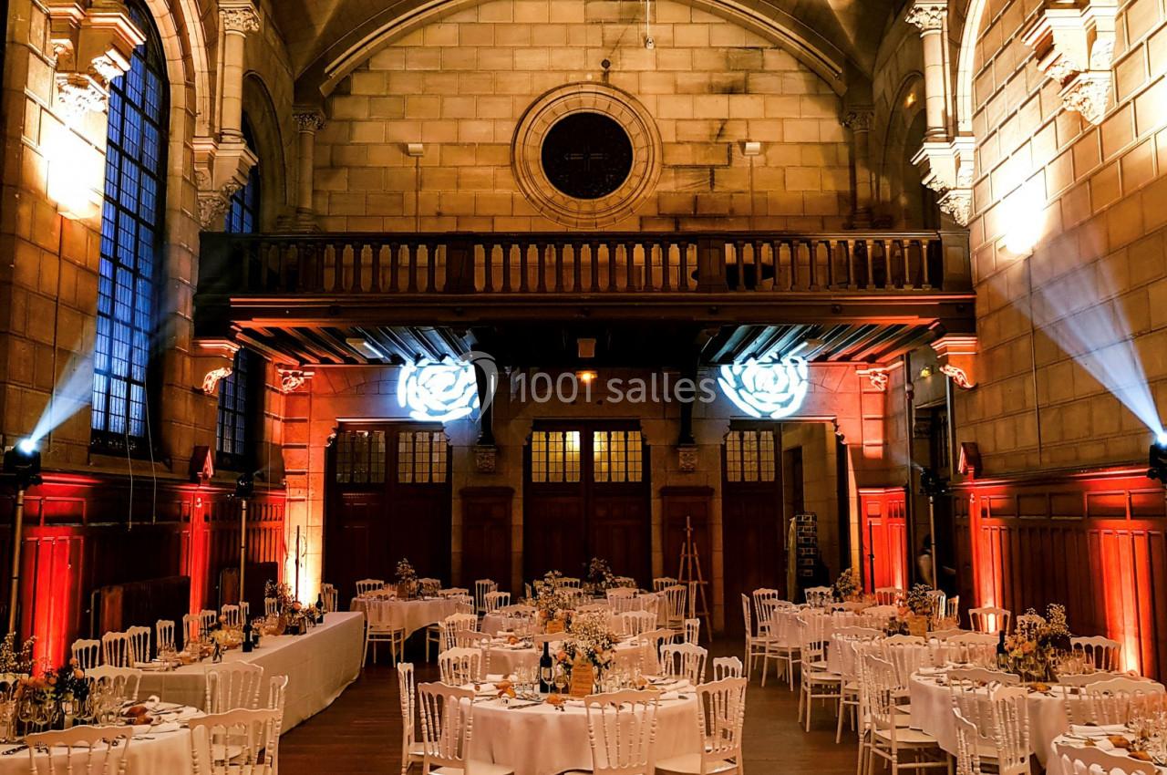 Salle de réception décorée avec des tables dressées, éclairage chaleureux et architecture en pierre avec vitraux.