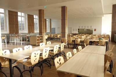 Salle lumineuse avec de grandes fenêtres, tables en bois clair et chaises alignées dans un espace de restauration collectif.