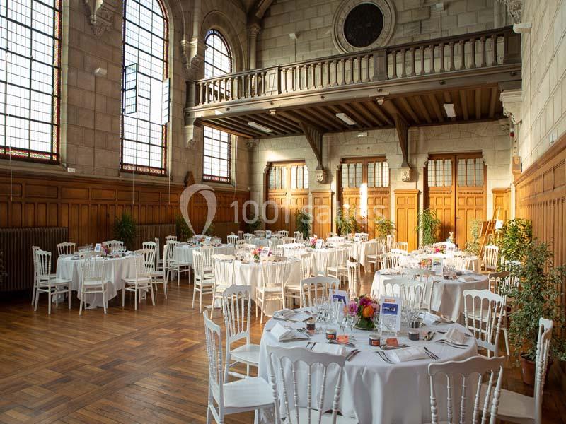 Salle de réception lumineuse avec des tables rondes dressées, des chaises blanches et des fenêtres en vitraux.