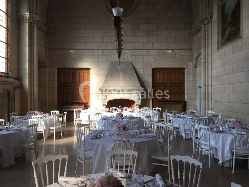 Salle de réception lumineuse avec des tables rondes dressées, des chaises blanches et une grande cheminée en pierre.