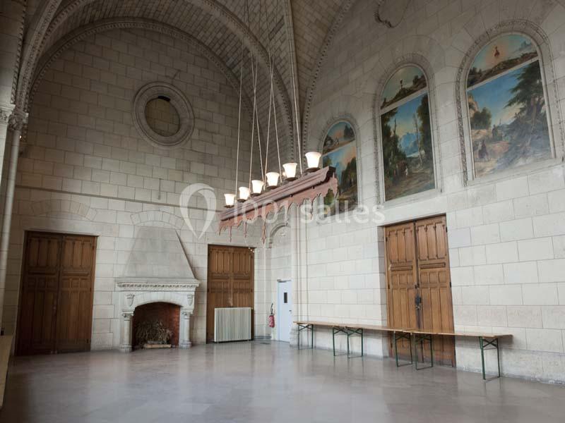 Grande salle avec des murs en pierre, une cheminée, des portes en bois, des fresques murales et un lustre suspendu.