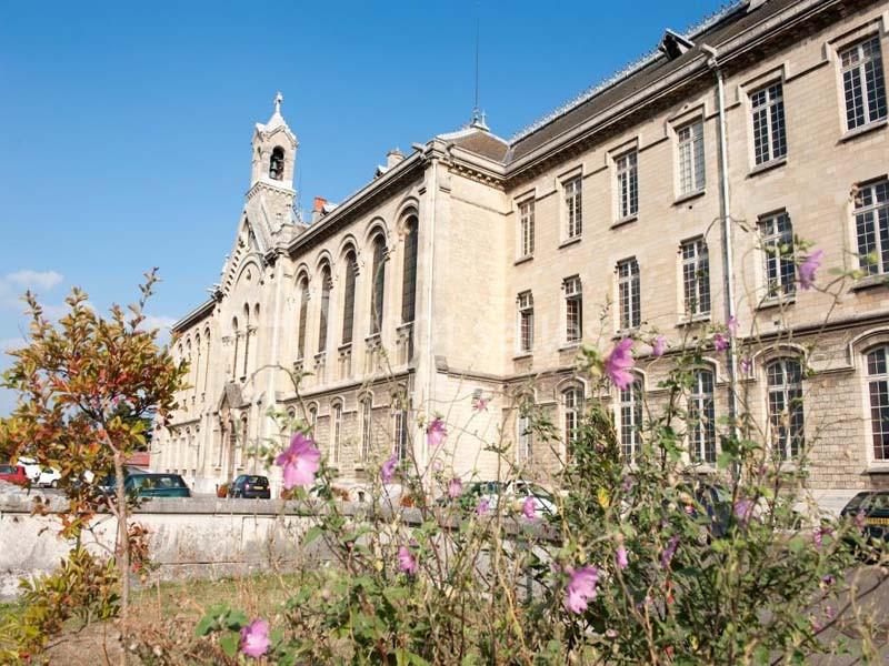 Façade d'un bâtiment ancien en pierre avec des fenêtres en arc, vue depuis un jardin fleuri au premier plan.