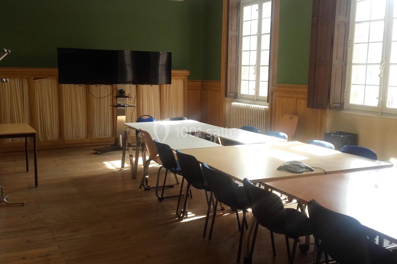 Salle de réunion lumineuse avec tables disposées en rectangle, chaises, écran mural et porte-manteau.