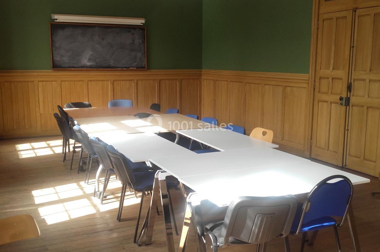 Salle de réunion lumineuse avec tables blanches disposées en rectangle, chaises variées et tableau noir au fond.