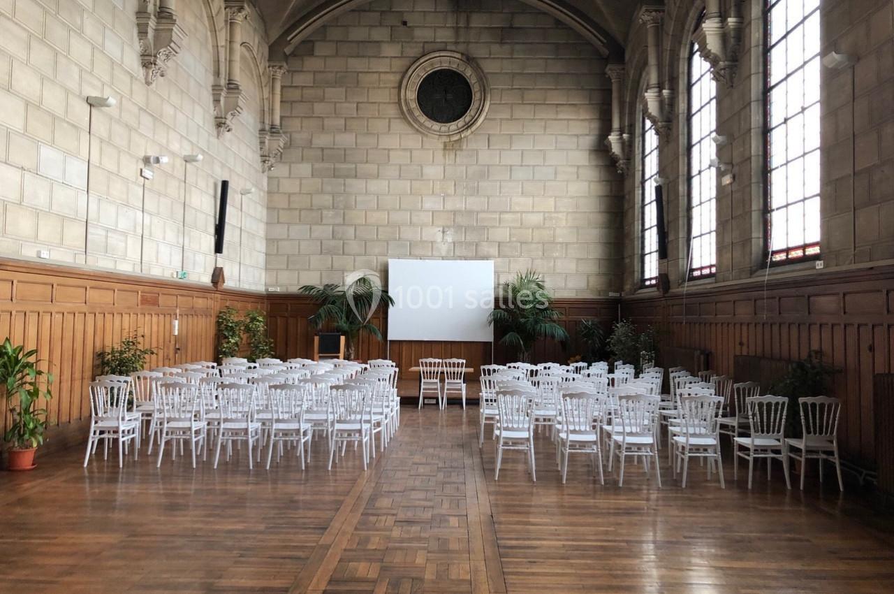 Salle avec parquet, murs en pierre, grandes fenêtres et chaises blanches disposées face à un écran de projection.
