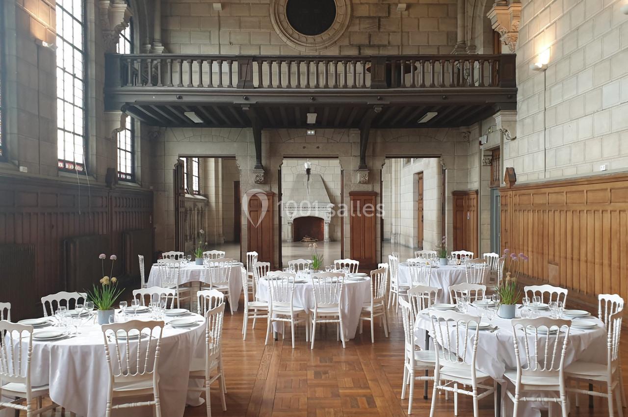 Salle de réception avec tables rondes dressées, nappes blanches et chaises, dans un décor en bois et pierre.