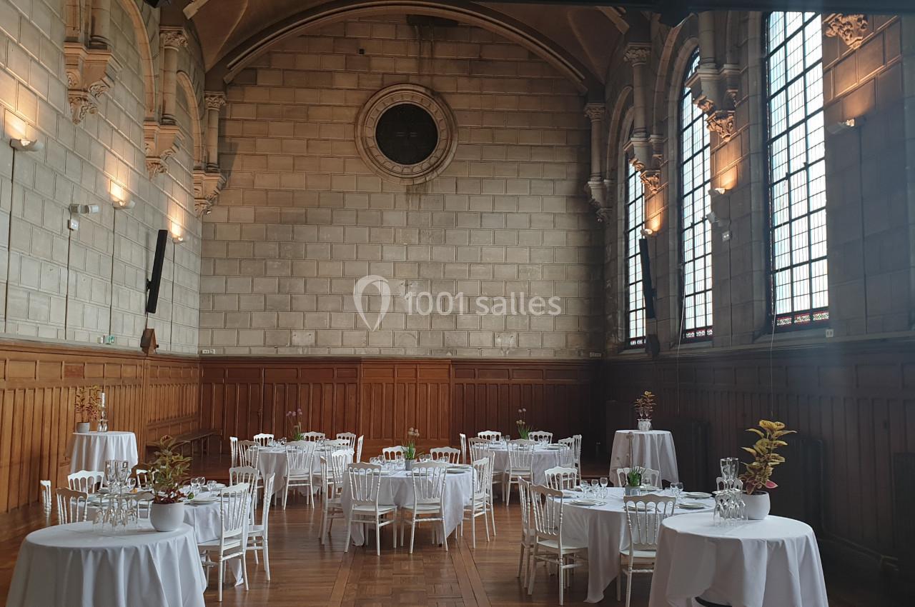 Salle de réception avec des tables rondes dressées, entourées de chaises blanches, dans un décor aux murs en pierre et…