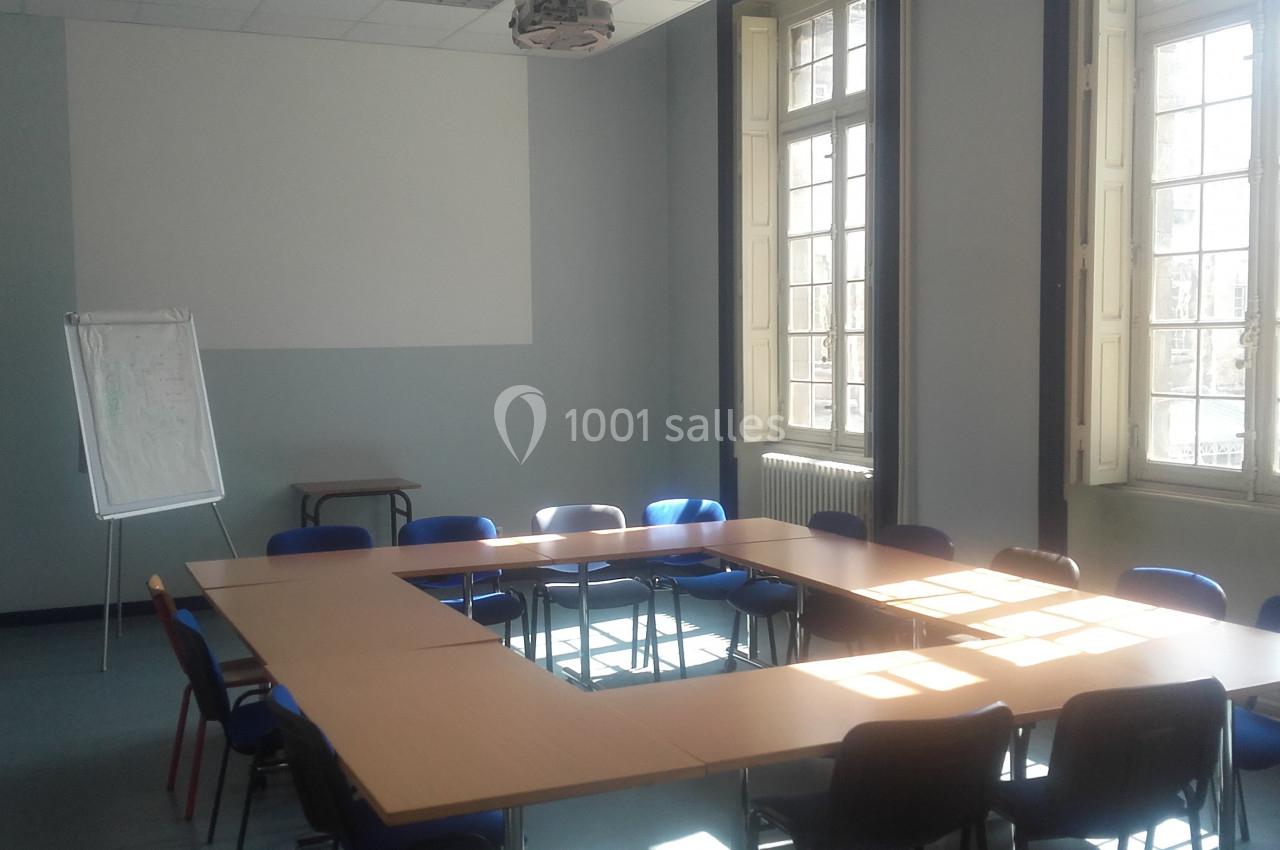 Salle de réunion lumineuse avec tables disposées en U, chaises bleues et un tableau de présentation sur pied.