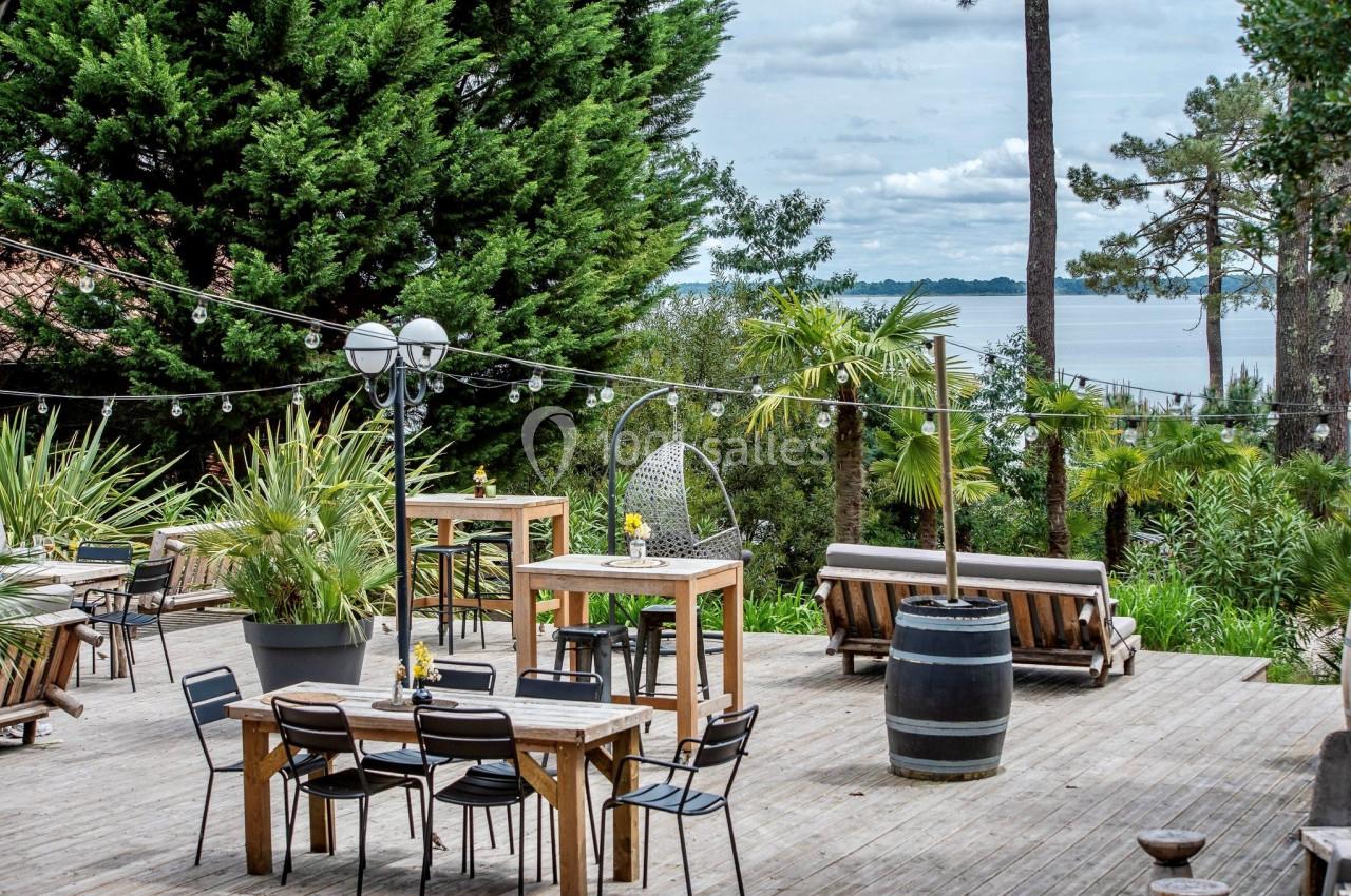 Terrasse en bois aménagée avec tables, chaises, plantes et vue sur un lac entouré de végétation.