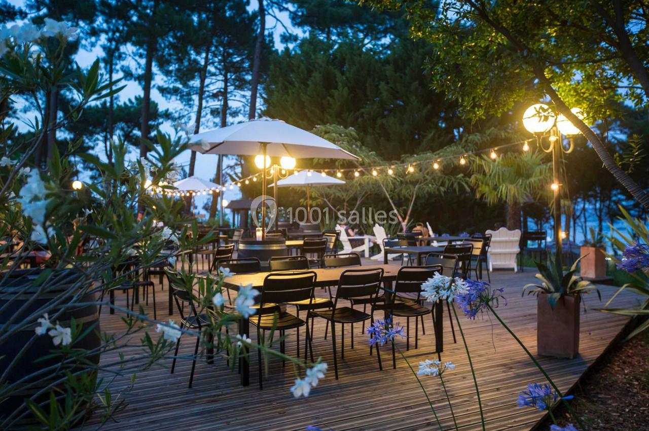 Terrasse en bois éclairée par des guirlandes lumineuses et lampadaires, entourée de végétation et de tables avec chaises.