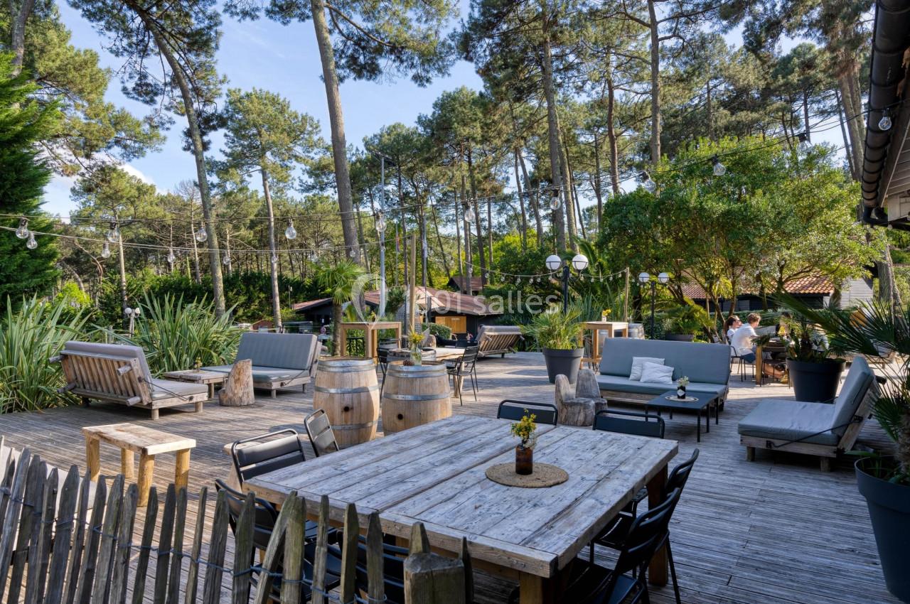 Terrasse en bois aménagée avec tables, fauteuils et plantes, entourée de pins sous un ciel dégagé.