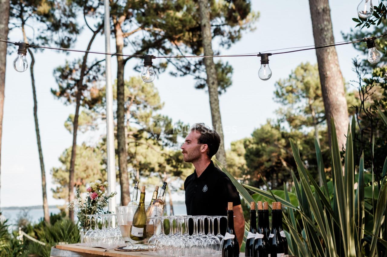 Un serveur derrière un bar extérieur fait de tonneaux, entouré de verres, bouteilles et végétation, sous des guirlandes…