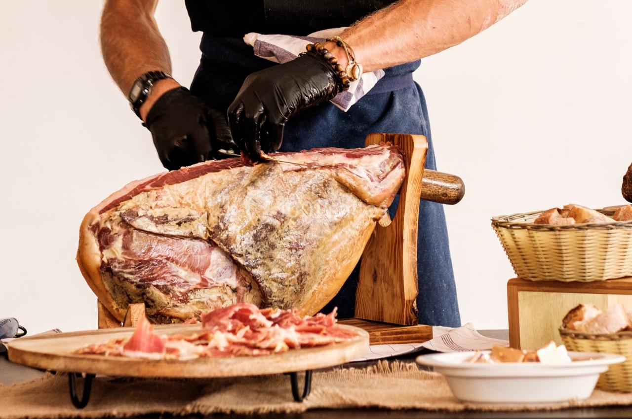 Un artisan découpe un jambon sec sur un support en bois, avec des tranches disposées sur une assiette au premier plan.