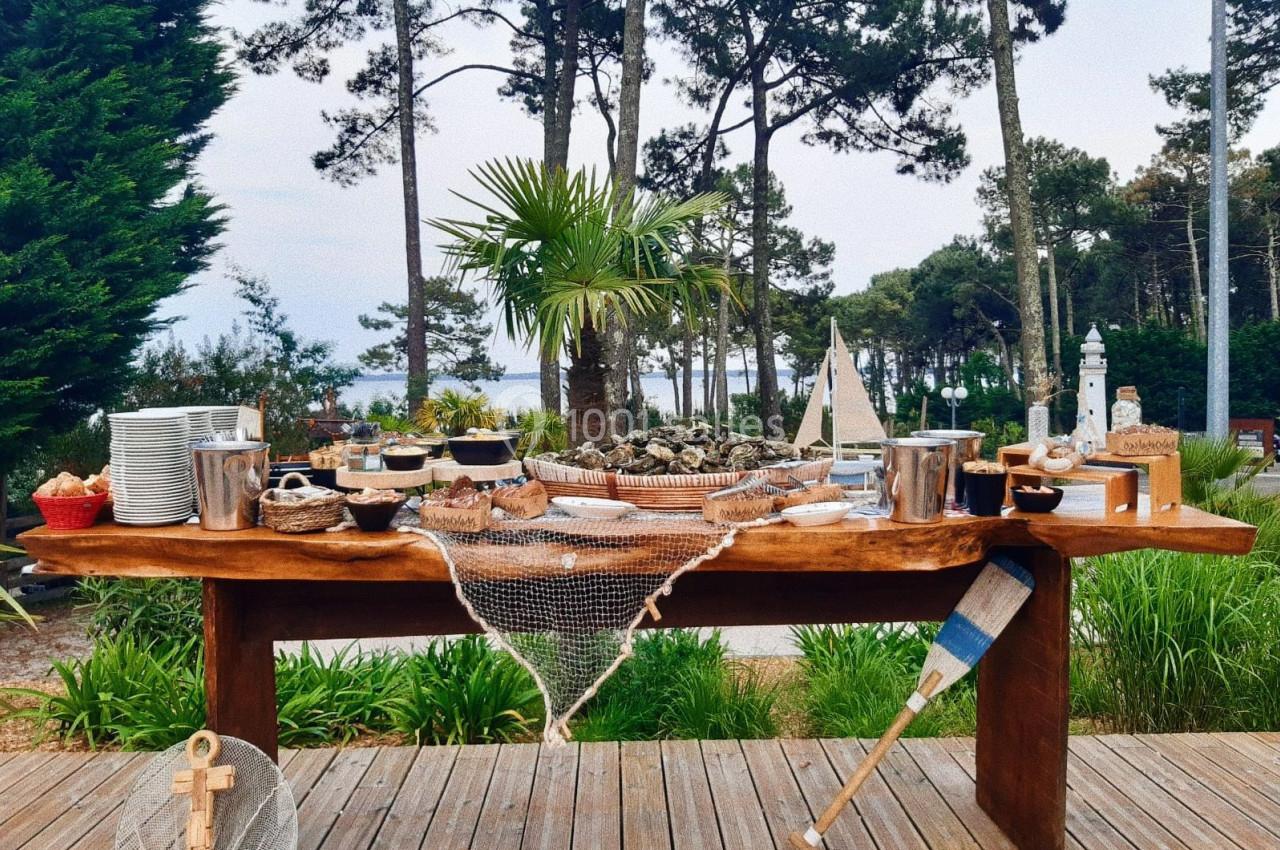 Table en bois garnie de fruits de mer, vaisselle et décorations maritimes, située en extérieur près d'une forêt.