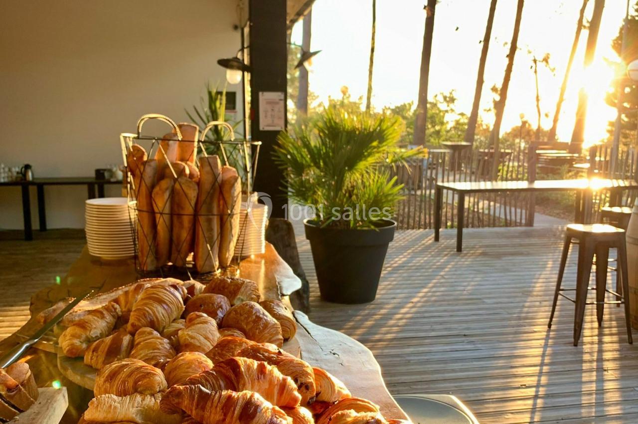 Corbeille de baguettes et plateau de croissants sur une table en bois, terrasse ensoleillée en arrière-plan.