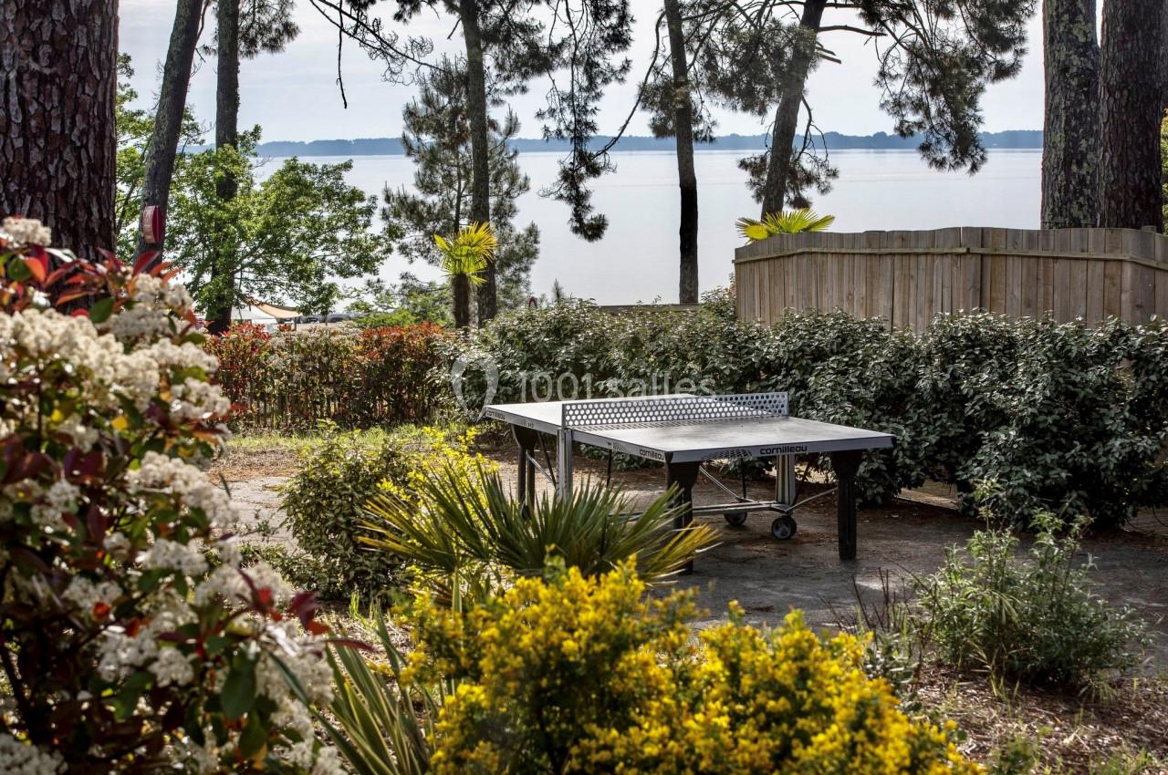 Table de ping-pong en extérieur, entourée de végétation, avec vue sur un plan d'eau et des arbres en arrière-plan.