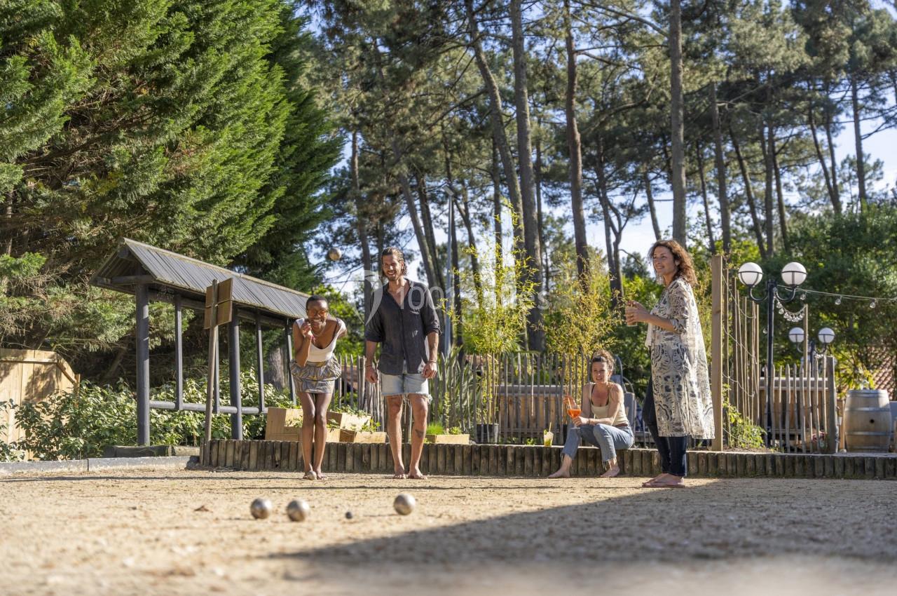 Des personnes jouent à la pétanque sur un terrain ensoleillé entouré d'arbres et de structures en bois.