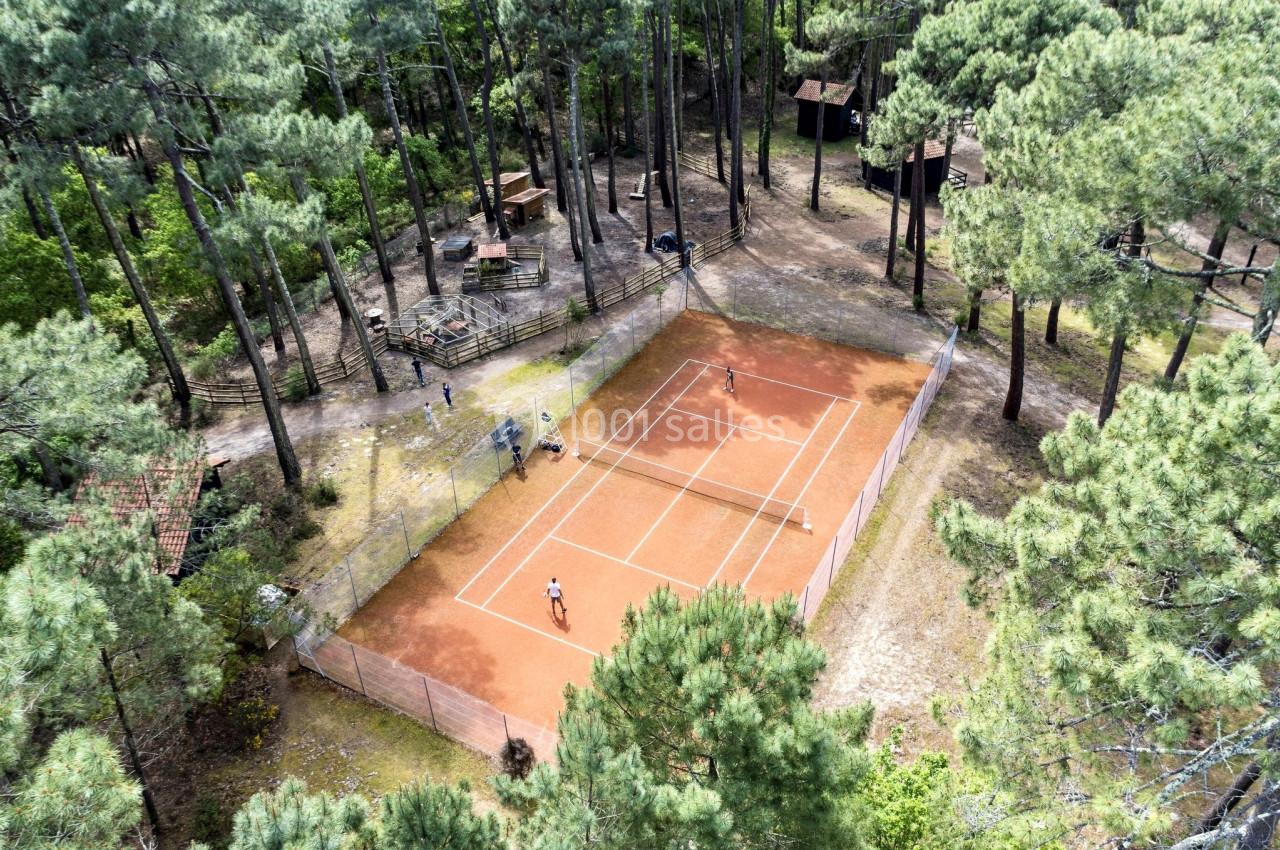 Vue aérienne d'un court de tennis en terre battue entouré de pins, avec deux joueurs en pleine partie.