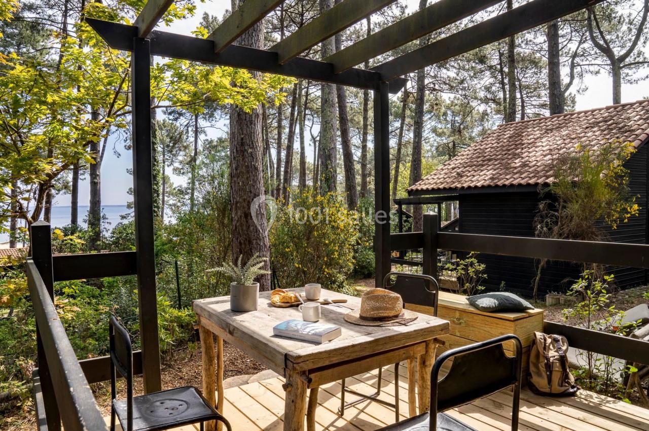 Table en bois dressée sur une terrasse ombragée, entourée de pins, avec vue sur un lac et une cabane en arrière-plan.
