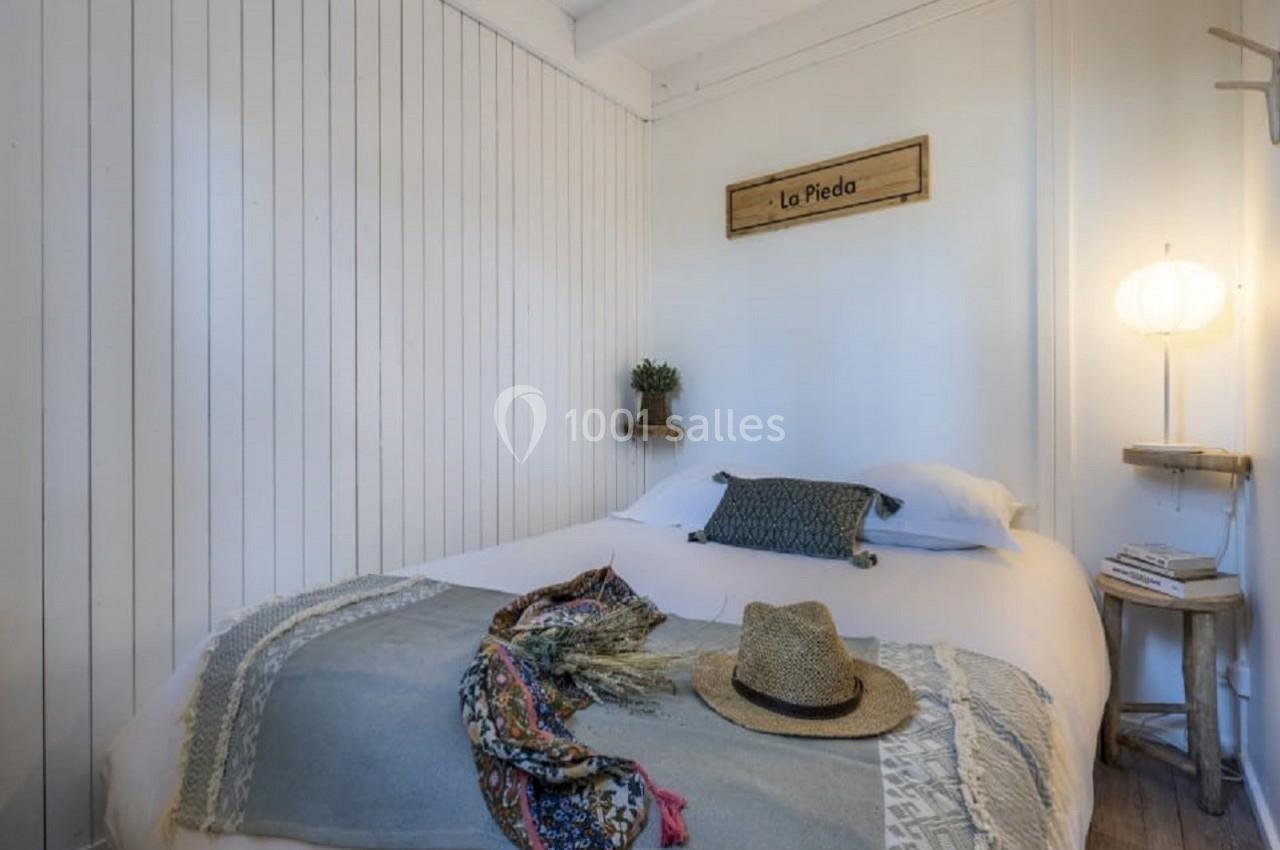 Lit double avec couvre-lit, chapeau et foulard posés dessus, dans une chambre lumineuse aux murs en bois blanc.