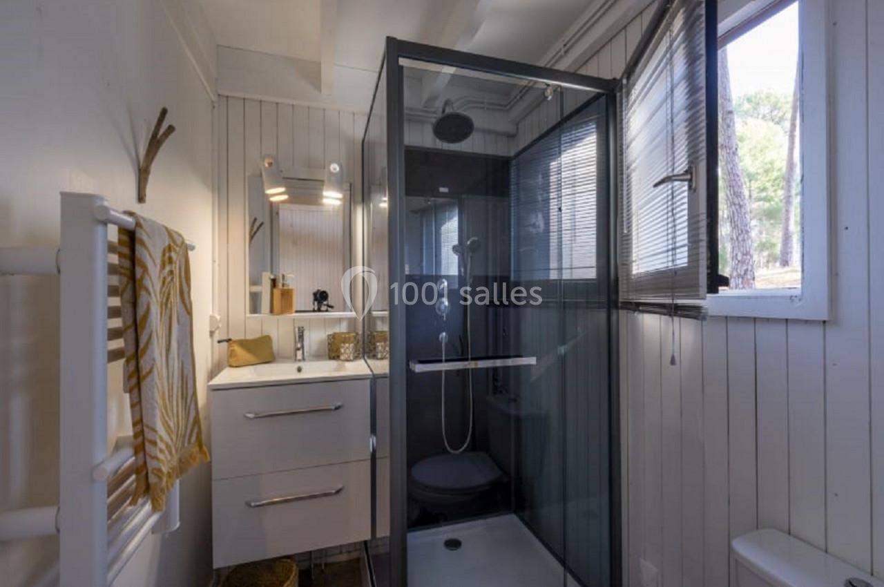 Salle de bain lumineuse avec cabine de douche vitrée, meuble vasque blanc et fenêtre laissant entrer la lumière naturelle.