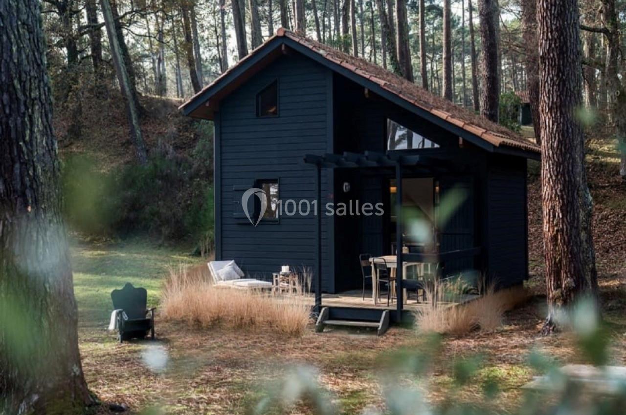 Cabane en bois sombre avec terrasse, entourée de pins et d'herbes hautes dans une clairière forestière.