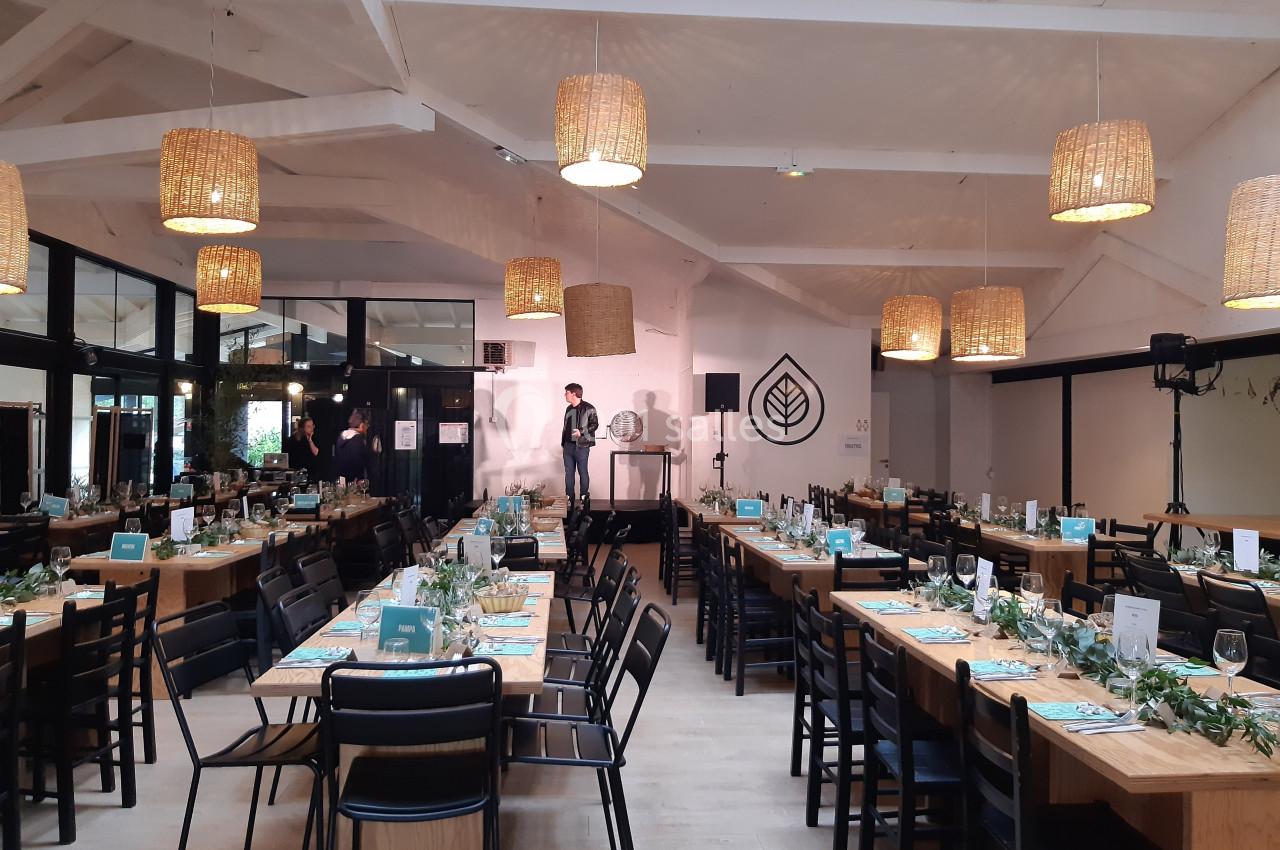 Salle de réception lumineuse avec tables dressées, décorations végétales et suspensions en osier.