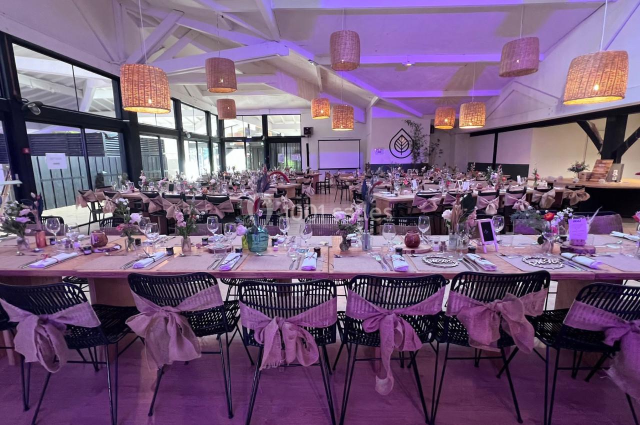 Salle de réception décorée avec des tables alignées, nappes en toile de jute, fleurs et éclairage violet.