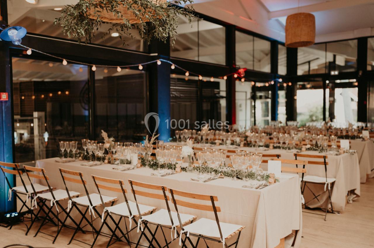 Salle de réception décorée avec des tables longues, nappes blanches, chaises en bois et guirlandes lumineuses suspendues.