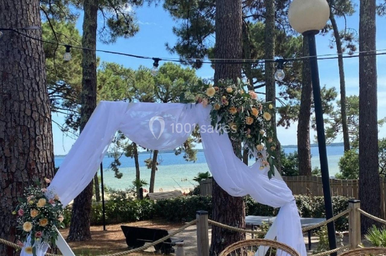 Arche décorée de voilages blancs et de fleurs, entourée de chaises en rotin, avec vue sur une forêt et la mer.