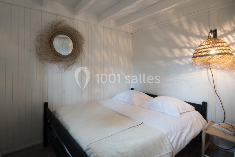 Lit double avec draps blancs dans une chambre lumineuse, décorée de bois clair, d'un miroir rond et d'une lampe en osier.