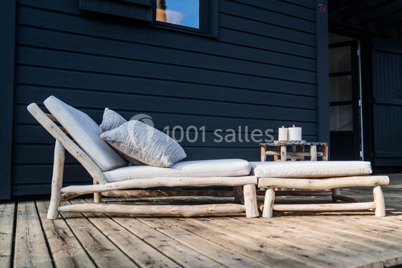Chaise longue en bois clair avec coussins blancs, posée sur une terrasse en bois devant un mur sombre.