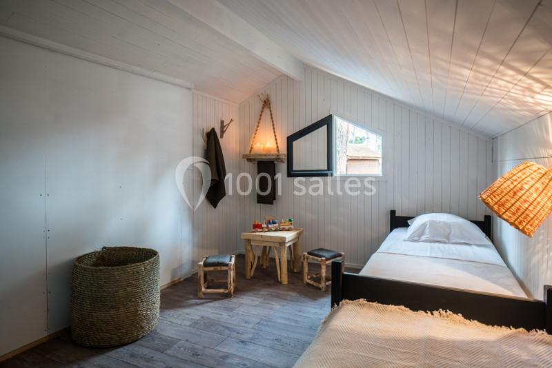 Chambre mansardée avec lit simple, table en bois, tabourets, panier tressé et lumière naturelle entrant par une fenêtre.