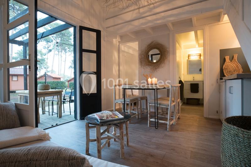 Intérieur d'une maison lumineuse avec salon, table à manger, bougies allumées et vue sur une terrasse extérieure boisée.