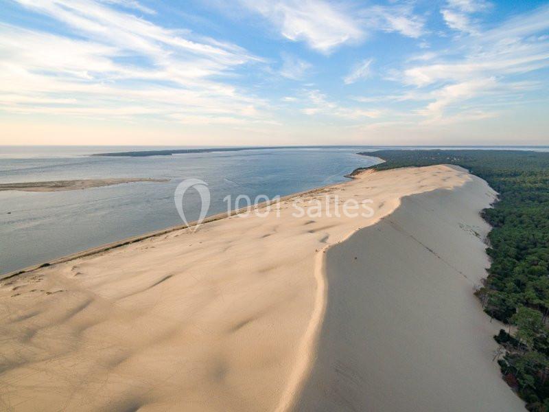 Vue aérienne d'une vaste dune de sable bordée par une forêt dense et un littoral sous un ciel partiellement nuageux.