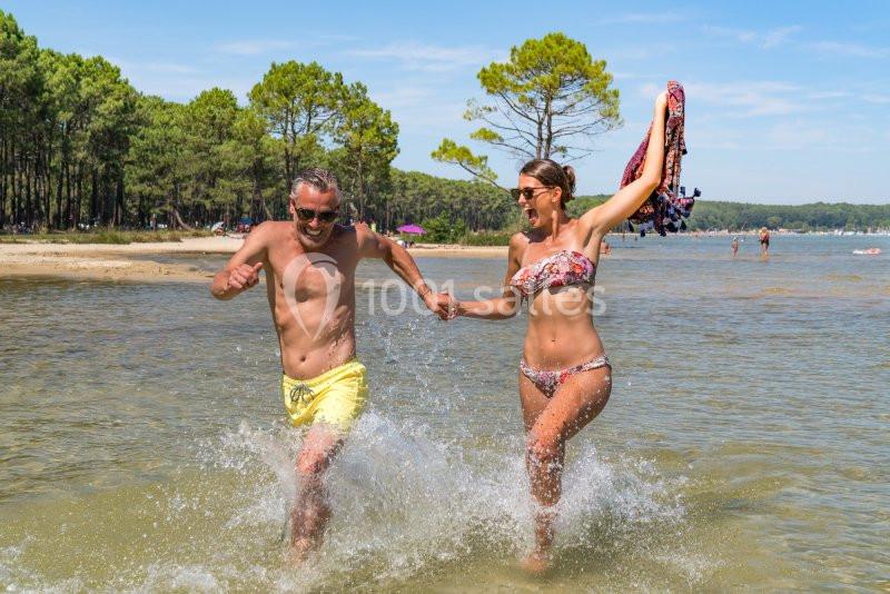 Un couple souriant court dans l'eau peu profonde d'un lac bordé de pins par une journée ensoleillée.