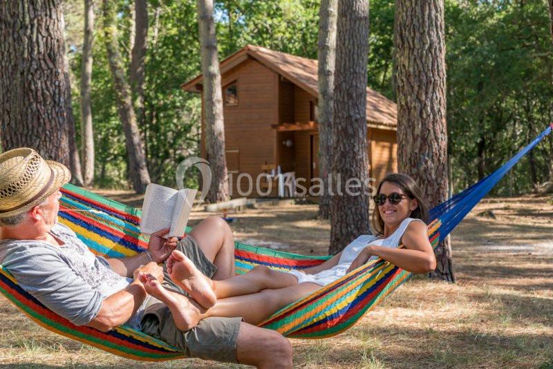 Deux personnes se détendent dans un hamac coloré, entourées d'arbres, avec une cabane en bois en arrière-plan.