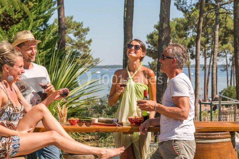Des amis discutent et rient autour d'une table en bois, avec des verres de vin, en plein air près d'une forêt et d'un lac.