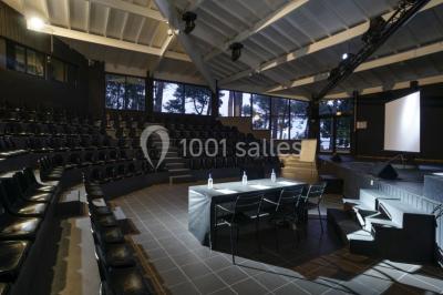 Cabane en bois sombre entourée de grands pins dans une forêt, avec lumière du soleil filtrant à travers les arbres.