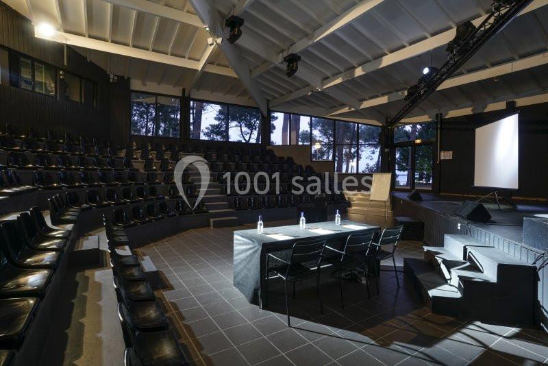 Salle de conférence moderne avec gradins, table de présentation, chaises et écran de projection, éclairée en soirée.