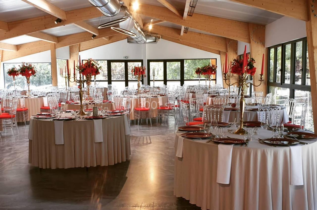 Salle de réception lumineuse avec tables rondes dressées, décorations florales rouges et chaises transparentes.