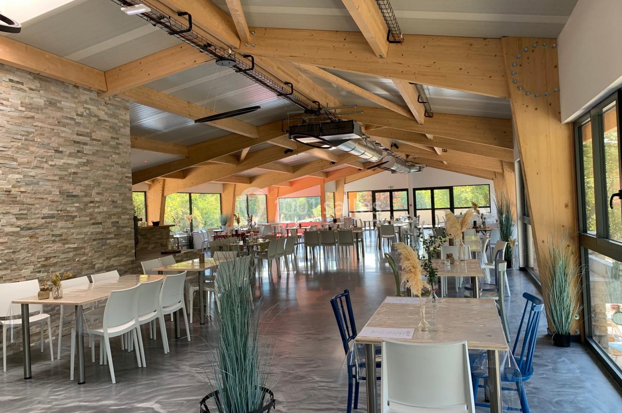 Salle lumineuse avec tables et chaises modernes, poutres en bois apparentes et grandes baies vitrées donnant sur l'extérieur.