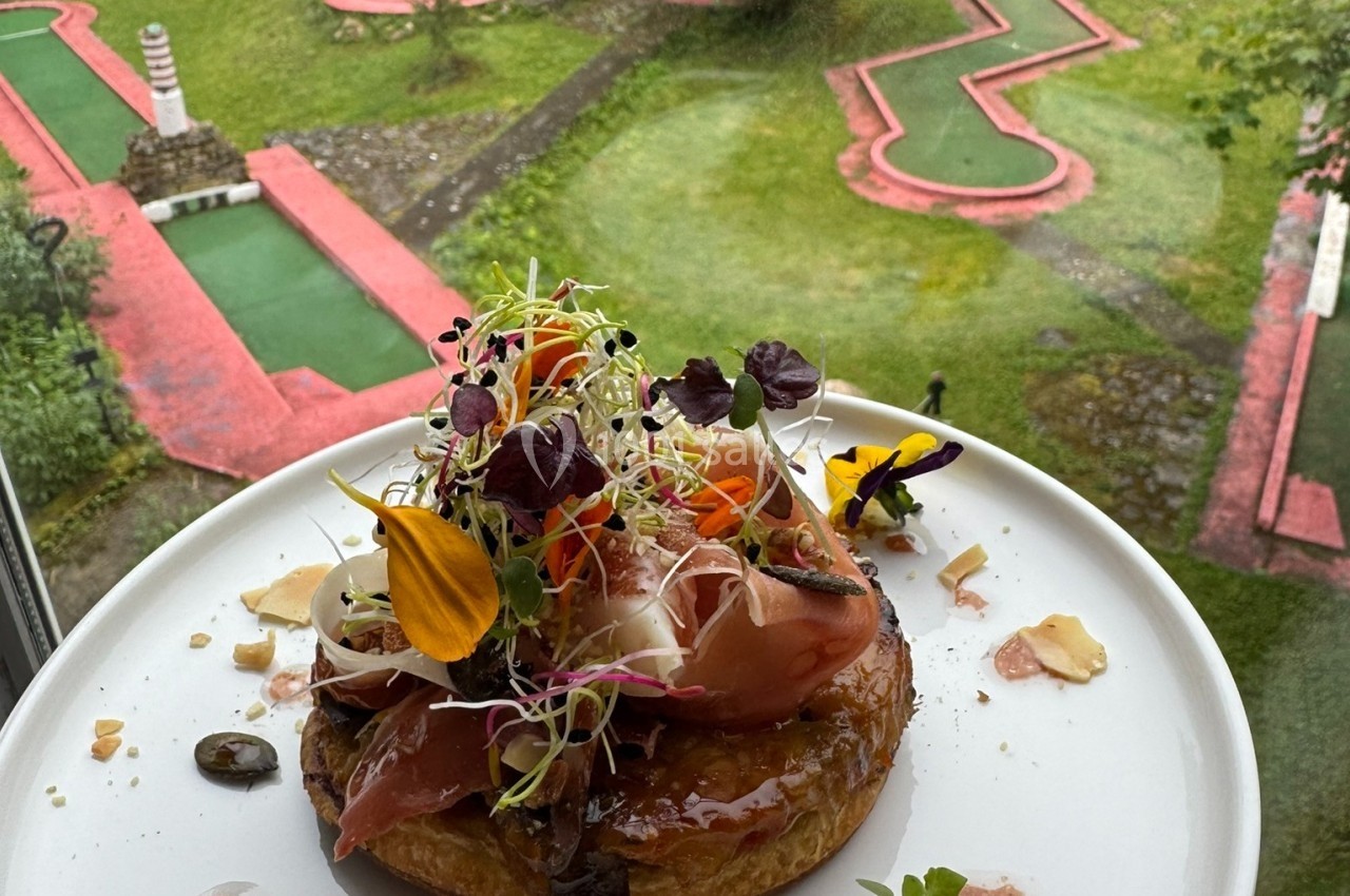 Tarte garnie de légumes, jambon et pousses, présentée devant un mini-golf verdoyant vu depuis une fenêtre.
