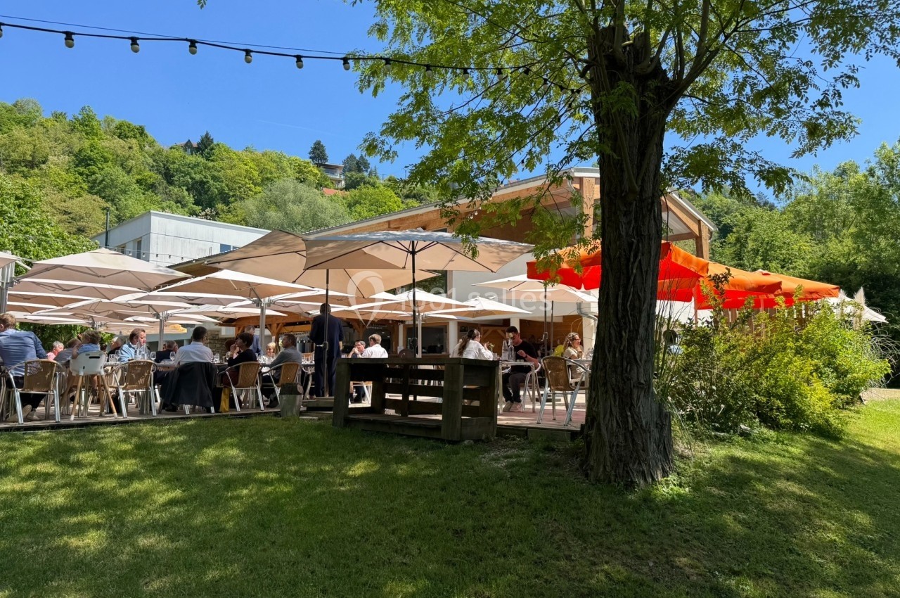 Terrasse d'un restaurant avec des tables ombragées par des parasols, entourée de verdure et d'arbres.