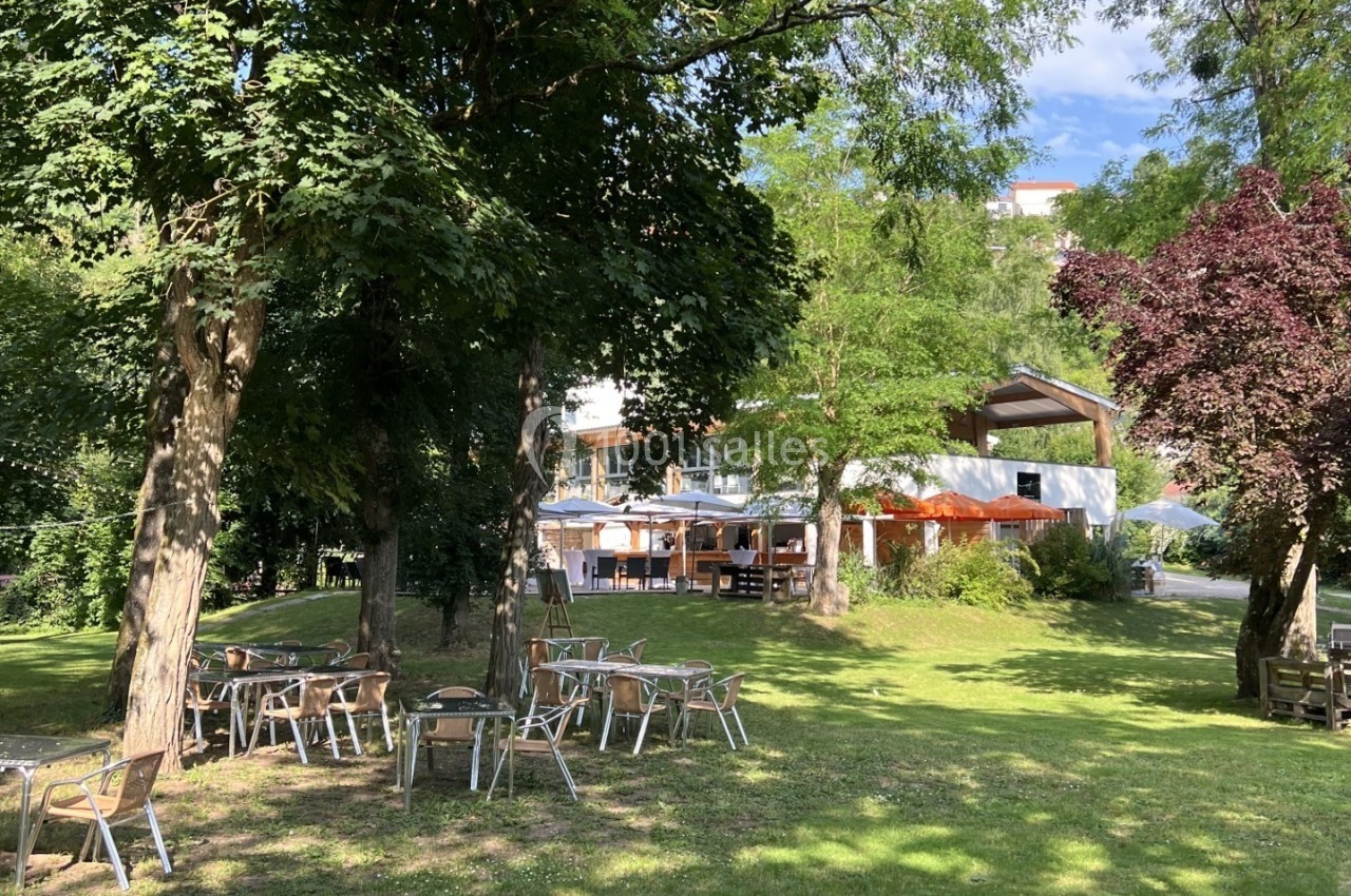 Chaises et tables disposées sur une pelouse ombragée par des arbres, avec un bâtiment en arrière-plan.